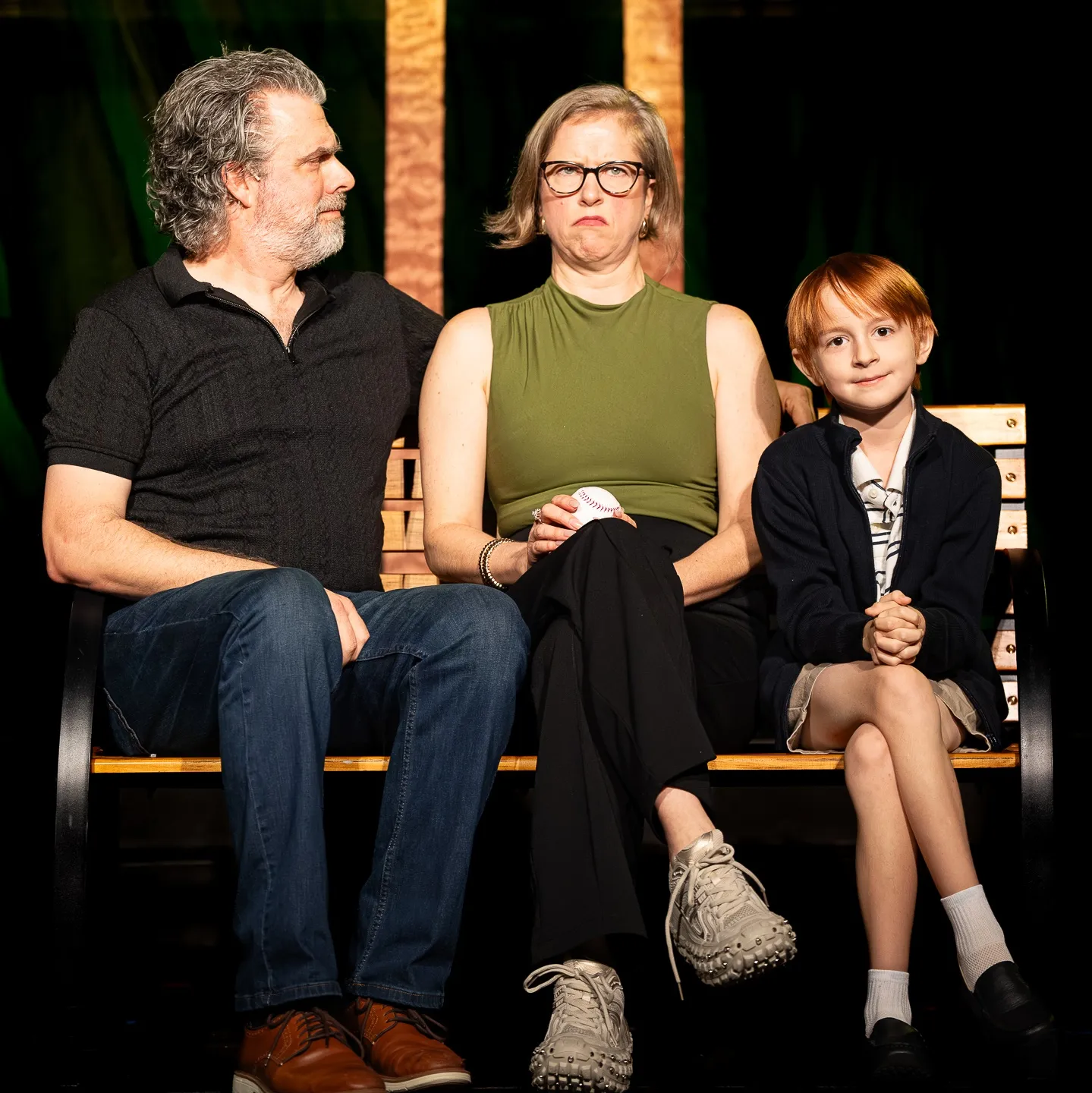 Three actors sit on a bench in a stage scene. Jeff Drushal looks toward Karen Konzen, who wears glasses and pulls a comically unimpressed face while holding a baseball, as young Flynn Linning smiles beside them.