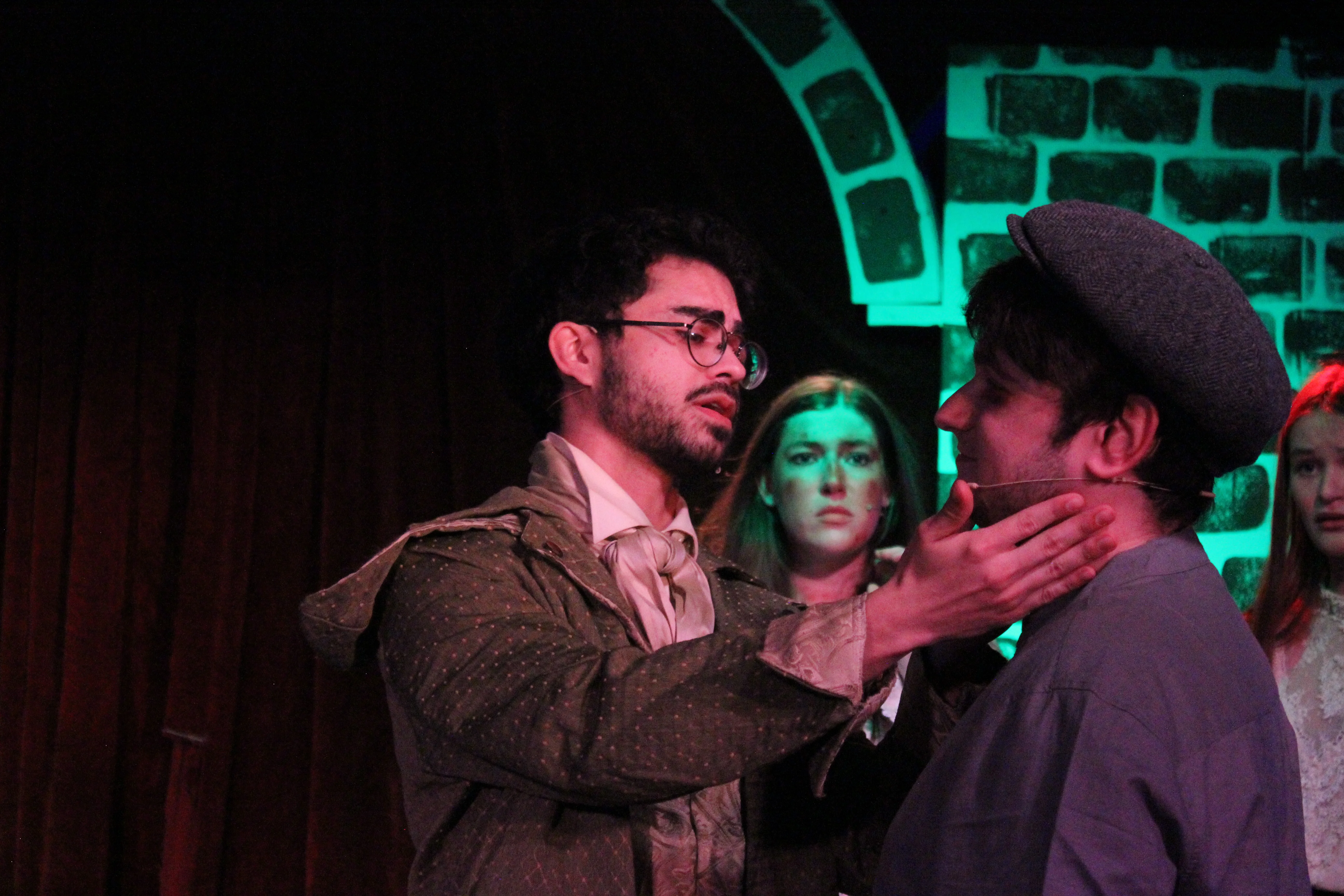 A costumed performer grips another performer's chin in a tense close-up from Swoop Troupe's Sweeney Todd, with ensemble members watching under green and red stage light.
