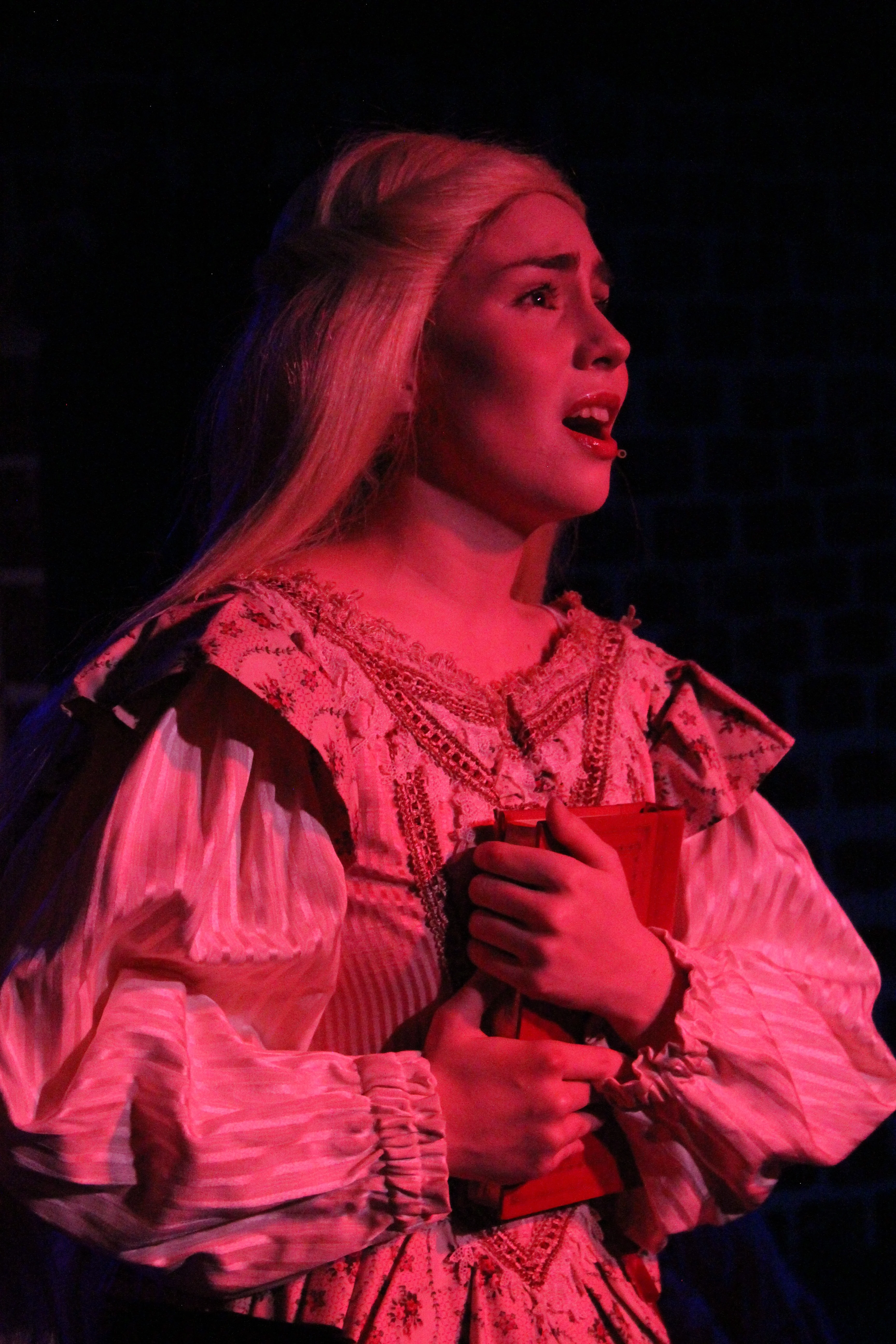 A blonde performer in a white period costume lies under red light, clutching a book to her chest during Swoop Troupe's Sweeney Todd.