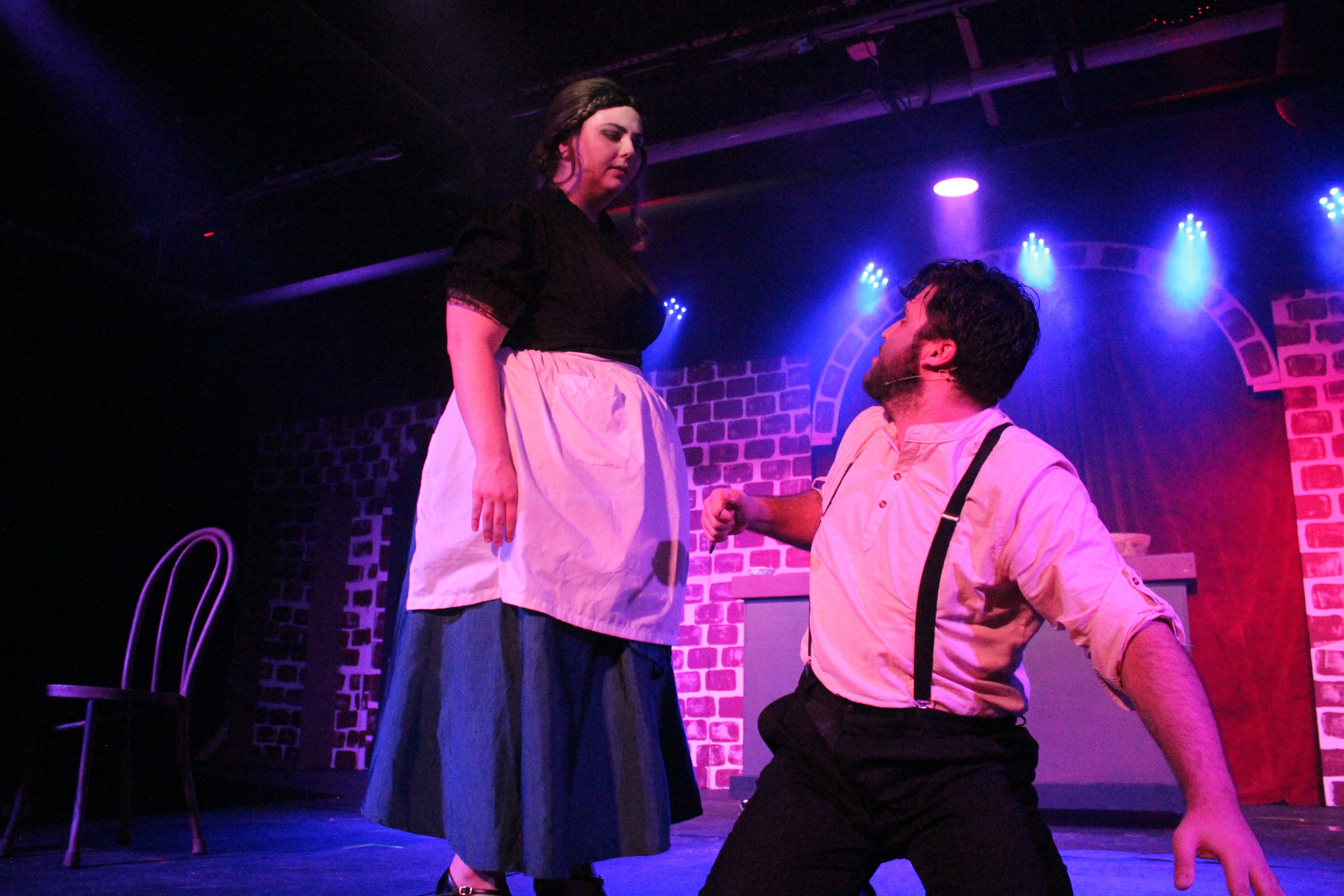 Mrs. Lovett stands over Sweeney Todd as he kneels beside the pie-shop counter beneath blue and magenta lights.